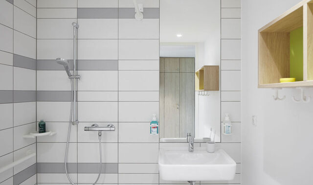 Badezimmer eines Patientenzimmers in der Klinik Königshof. In diesem befindet sich eine Dusche, ein Waschbecken mit einem Spiegel über diesem. Außerdem hängt ein kleines Regal an der Wand.