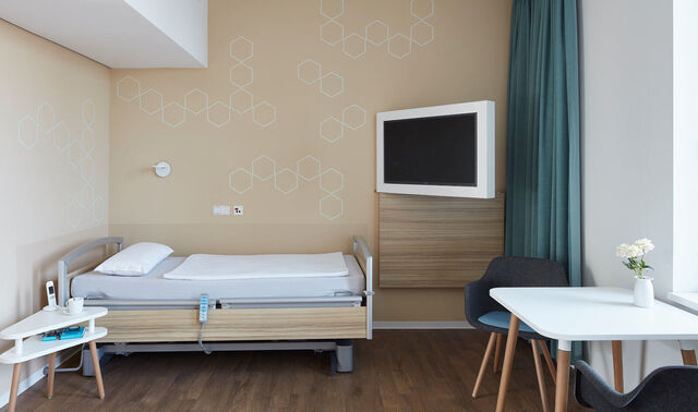 Patientenzimmer mit einem Bett, einem Nachttisch, einem Schreibtisch und einem Fernseher.