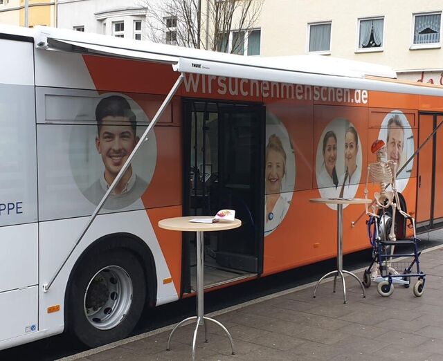 Ein Bus, welcher die Werbung für die St. Augustinus Gruppe trägt. Vor diesem stehen zwei Stehtisch und ein Skelette mit einem Rollator. 