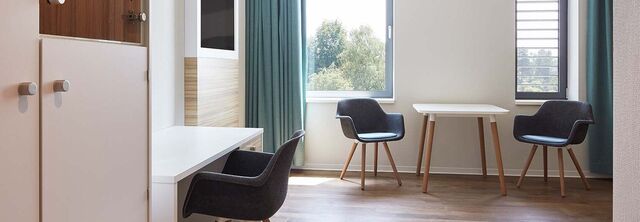 Ein Patientenzimmer in dem sich einige Stühle sowie einen Schreibtisch, einen Schrank und einen Fernseher befinden.