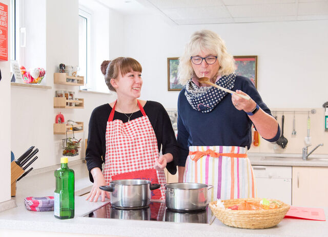 Zwei Frauen stehen an einem Herd auf welchem zwei Töpfe stehen. Sie tragen Schürzen und kochen etwas. Die eine Frau kostet das Essen mit einem langen Holzlöffel.