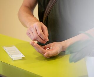 Eine Hand mit einigen Tabletten. Vor dieser liegt eine Schachtel für Tabletten auf einem Tisch.