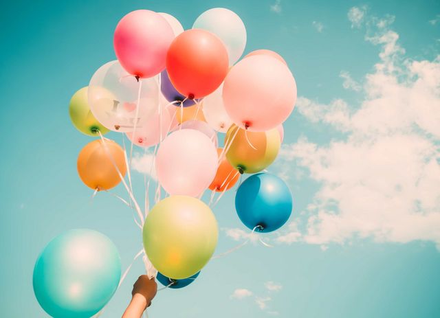 Eine Hand hält viele Ballons in den blauen Himmel. An diesem befindet sich eine kleine Wolke.